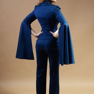 May include: A woman wearing a dark blue pantsuit with wide bell sleeves. The suit is tailored and has a fitted silhouette.