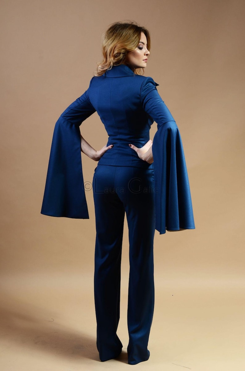 May include: A woman wearing a dark blue pantsuit with wide bell sleeves. The suit is tailored and has a fitted silhouette.