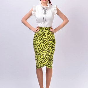 May include: A woman wearing a white blouse with a black bow and a green and black zebra print skirt. She is standing with her hands on her hips and looking at the camera.