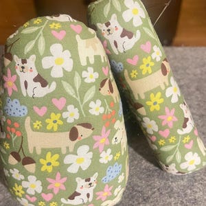 May include: Two fabric objects with a green background and a repeating pattern of cartoon dogs, flowers, hearts, and acorns. The dogs are in various poses, and the flowers are white, pink, and yellow. One object is cylindrical, and the other is rounded.