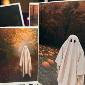 May include: Several Halloween-themed art prints featuring a ghost figure in a sheet. One print shows the ghost on a path in a forest, another shows the ghost in a pumpkin patch. The prints have a warm color palette.