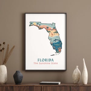 Puede incluir: Lámina artística enmarcada con una colorida ilustración del estado de Florida. La obra representa una escena de playa con palmeras, un velero y un flamenco. El texto "FLORIDA The Sunshine State" está en la parte inferior.