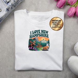 May include: White t-shirt featuring the text "I Love New Hampshire" above a scenic illustration. The design includes a moose, mountains, and pancakes. A gold sticker indicates premium quality and high resolution.