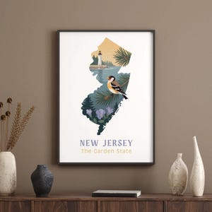 May include: Framed art print featuring the outline of New Jersey filled with illustrations of a lighthouse, bird, and flowers. The text "NEW JERSEY The Garden State" is printed at the bottom in a serif font. The artwork is displayed on a wooden surface.