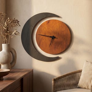 May include: A wall clock featuring a circular wooden face with a warm brown grain, set within a dark gray crescent moon frame. The clock has thin black hands and no visible numerals, creating a clean, geometric design that hangs on a wall.