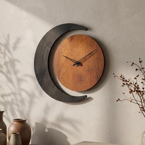 May include: A wall clock featuring a circular wooden face with a warm brown grain pattern, surrounded by a dark charcoal-colored crescent moon frame. The clock has two thin black hands positioned at the center of the circular wooden dial.