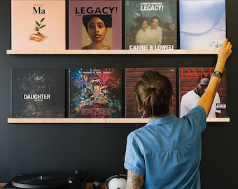 May include: A display of vinyl records on wooden shelves against a dark wall. The records feature album art with text including "Ma", "LEGACY!", "Carrie & Lowell", "DAUGHTER", and "Stranger Things". A turntable and speakers are visible below.