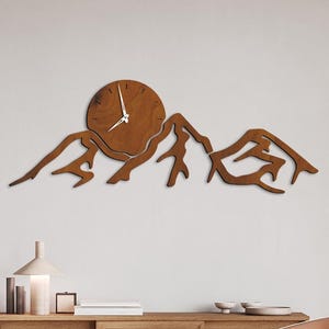 May include: A wooden wall clock in the shape of a mountain range. The clock face is round with hands indicating the time. The clock is made of brown wood and is mounted on a white wall. The color is Beech.