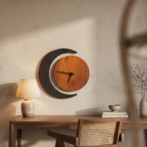 May include: A wall clock featuring a circular wooden face with a warm brown grain and black clock hands, partially framed by a dark, crescent-shaped wooden element. The clock is mounted on a plain wall above a wooden desk with a lamp and books.