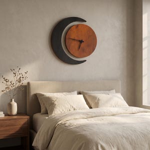 May include: A wall clock featuring a circular wooden face in a warm brown tone, partially framed by a dark charcoal crescent moon shape. The clock has thin black hands positioned at the center of the circular face, mounted on a plain wall.