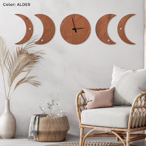 May include: A wooden wall clock in the shape of the phases of the moon. The clock is alder-colored and has black clock hands. The design includes a full moon clock face and crescent moon shapes. The clock is mounted on a white wall.