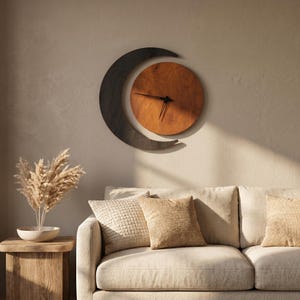 May include: A wall clock featuring a circular wooden face in a warm brown tone, partially enclosed by a dark charcoal-colored crescent moon shape. The clock has thin black hands and is mounted on a wall above a sofa with textured beige pillows.