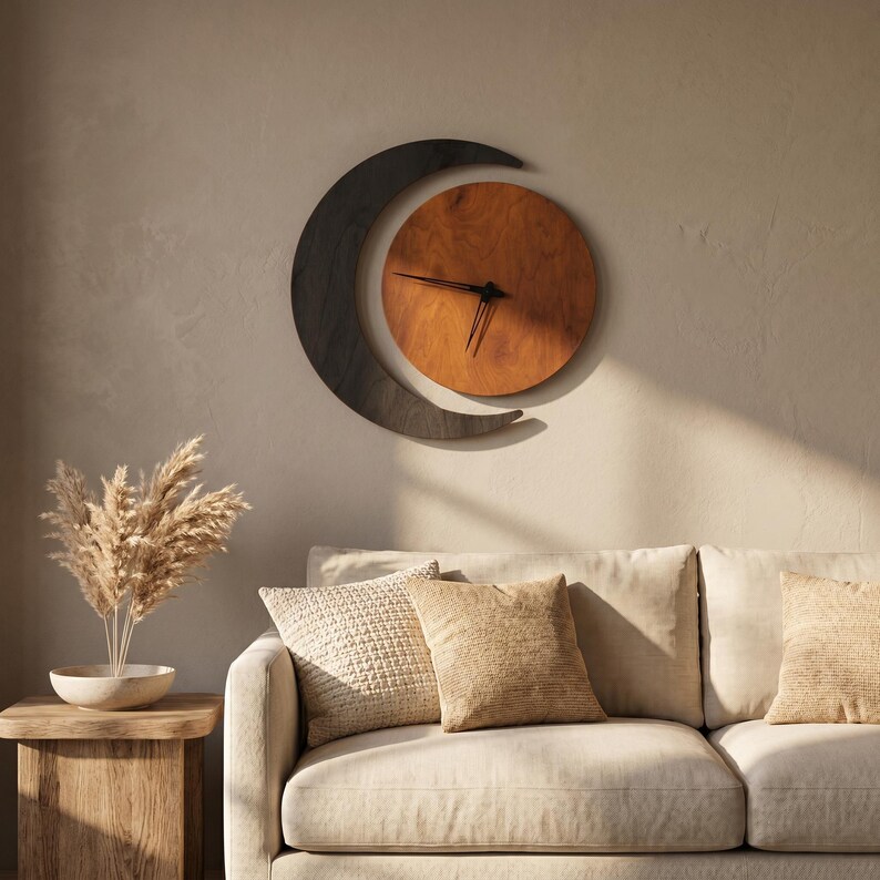 May include: A wall clock featuring a circular wooden face in a warm brown tone, partially enclosed by a dark charcoal-colored crescent moon shape. The clock has thin black hands and is mounted on a wall above a sofa with textured beige pillows.