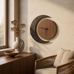 May include: A wall clock featuring a circular wooden face with black hands, partially framed by a dark brown crescent moon shape. The clock is mounted on a wall, showcasing a natural wood grain finish and a clean, geometric design.
