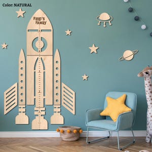 May include: A wooden rocket-shaped growth chart with the text "FIELD'S FAMILY" and measurement markings. The natural wood-colored chart is mounted on a teal wall, surrounded by wooden star, planet, and UFO decorations. A light blue armchair with a yellow star pillow and a giraffe toy are in the foreground.