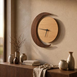 May include: A circular wall clock featuring a light oak wood face and a dark walnut wood crescent moon frame. The clock has two thin black metal hands positioned at the center. The design combines a circular clock face with a crescent shape.