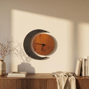 May include: A wall clock featuring a circular wooden face with a warm brown grain, partially encircled by a dark, crescent-shaped frame. The clock has thin black hands positioned at the center of the wooden circle, mounted on a plain wall.