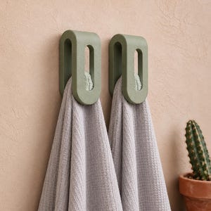 May include: Two sage green, U-shaped wall hooks mounted side-by-side. Each hook features a smooth, rounded rectangular frame with a central opening. A light gray, textured waffle-weave towel hangs through the center of each individual hook.