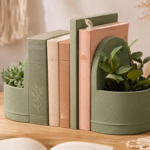 May include: A set of decorative bookends in a sage green color, holding a row of books in shades of green, peach, and cream. One bookend features a built-in planter with a succulent, and another with a leafy plant.