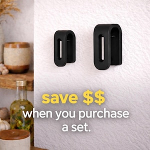 May include: Two black, U-shaped wall-mounted hooks of different sizes are attached to a textured white surface. The text "save $$ when you purchase a set." is displayed in yellow and white font below the hooks, indicating a bundle offer.