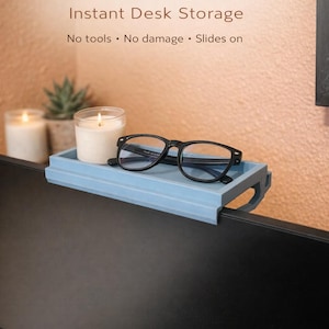 Op de afbeelding: Een lichtblauwe monitorplank met de tekst "Monitor Top Shelf Instant Desk Storage". De plank bevat een bril met zwart montuur, een brandende kaars en een kleine vetplant. De plank schuift op de monitor.