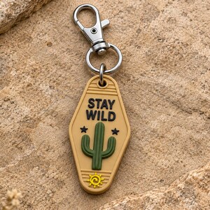 May include: A tan, motel-style keychain featuring the text STAY WILD in black, a green cactus graphic, two small black stars, and a yellow sun icon at the bottom. The keychain is attached to a silver-toned metal lobster claw clasp and ring.