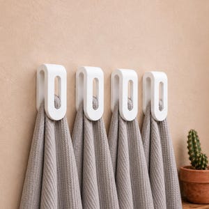 May include: Four white, U-shaped wall hooks mounted in a horizontal row, each holding a folded, light gray, textured waffle-weave towel. The hooks have a smooth, rounded design with a central vertical opening for hanging items securely.
