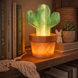 May include: A geometric green cactus lamp in a brown pot sits on a wooden shelf. The lamp is lit from within, casting a warm glow. Books and decorative items are in the background.