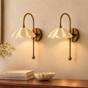 Op de afbeelding: Een paar wandlampen met gebogen, messingkleurige metalen armen en gebroken witte, golvende, klokvormige kappen. Elke lamp heeft een ronde muurplaat en een naar beneden gerichte stang die eindigt in een kleine bolvormige knop.