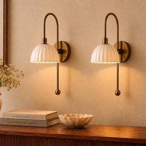 May include: A pair of wall-mounted lamps featuring curved brass-toned metal arms and white fluted, scalloped-edge shades. Each lamp has a circular wall plate and a vertical rod extending downwards with a small spherical finial at the bottom end.