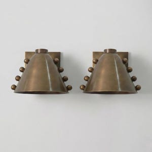 May include: Two bronze-colored wall sconces. Each sconce has a conical shade with a series of small, spherical accents. The sconces are mounted on square backplates. The design is symmetrical and modern.
