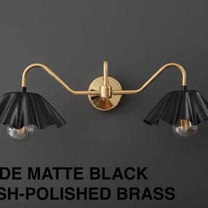 May include: A matte black and polished brass wall-mounted light fixture. The fixture has two black lampshades with a ruffled design, each housing a clear lightbulb. The text at the bottom reads "SHADE MATTE BLACK FINISH-POLISHED BRASS."