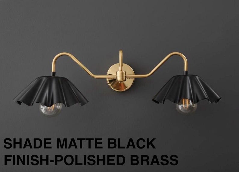 May include: A matte black and polished brass wall-mounted light fixture. The fixture has two black lampshades with a ruffled design, each housing a clear lightbulb. The text at the bottom reads "SHADE MATTE BLACK FINISH-POLISHED BRASS."
