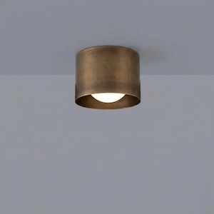 May include: A cylindrical flush mount ceiling light fixture with a brushed antique brass finish. The fixture features a solid metal exterior housing a single circular light source that emits a soft, warm glow from the center of the drum shade.