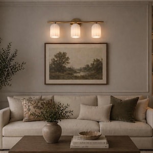May include: A living room scene featuring a cream-colored sofa with decorative pillows, a framed landscape painting, and a three-light fixture. A potted olive tree and a wooden coffee table complete the decor.
