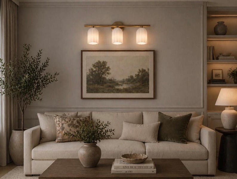 May include: A living room scene featuring a cream-colored sofa with decorative pillows, a framed landscape painting, and a three-light fixture. A potted olive tree and a wooden coffee table complete the decor.
