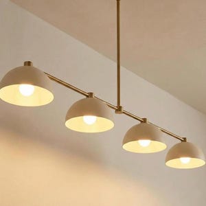 May include: A linear chandelier featuring a thin, straight brass-toned metal rod suspended from the ceiling. Four matte beige, dome-shaped shades are attached to the rod, each housing a single glowing light bulb that emits a warm, soft light.