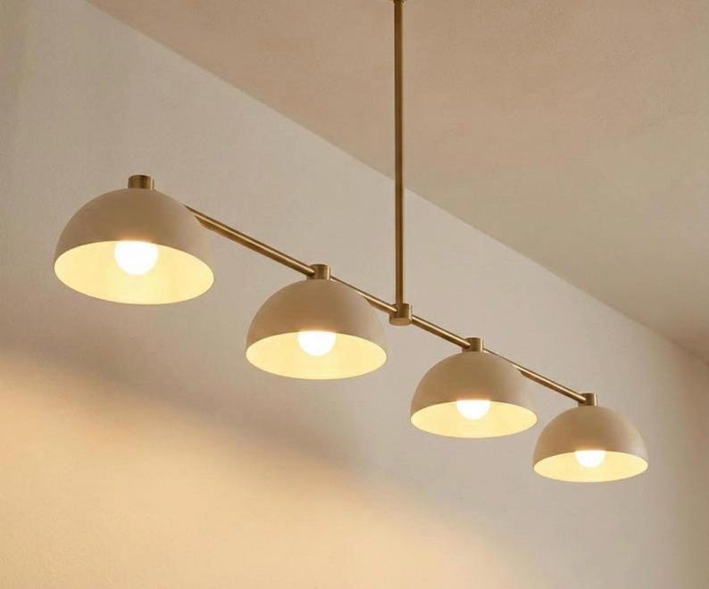 May include: A linear chandelier featuring a thin, straight brass-toned metal rod suspended from the ceiling. Four matte beige, dome-shaped shades are attached to the rod, each housing a single glowing light bulb that emits a warm, soft light.