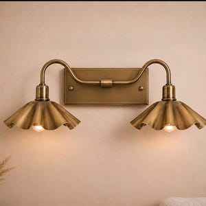 May include: A double wall sconce light fixture with a brushed brass finish. It features a rectangular backplate, two curved arms, and two downward-facing, fluted, scalloped-edge metal shades. Each shade houses a single glowing light bulb.