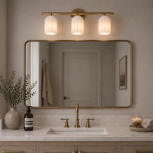 May include: A bathroom vanity with a gold-framed mirror, a three-light fixture, and a marble countertop. The vanity features a sink, faucet, and a decorative vase with greenery. A towel ring and a tray with candles and rolled towels are also visible.