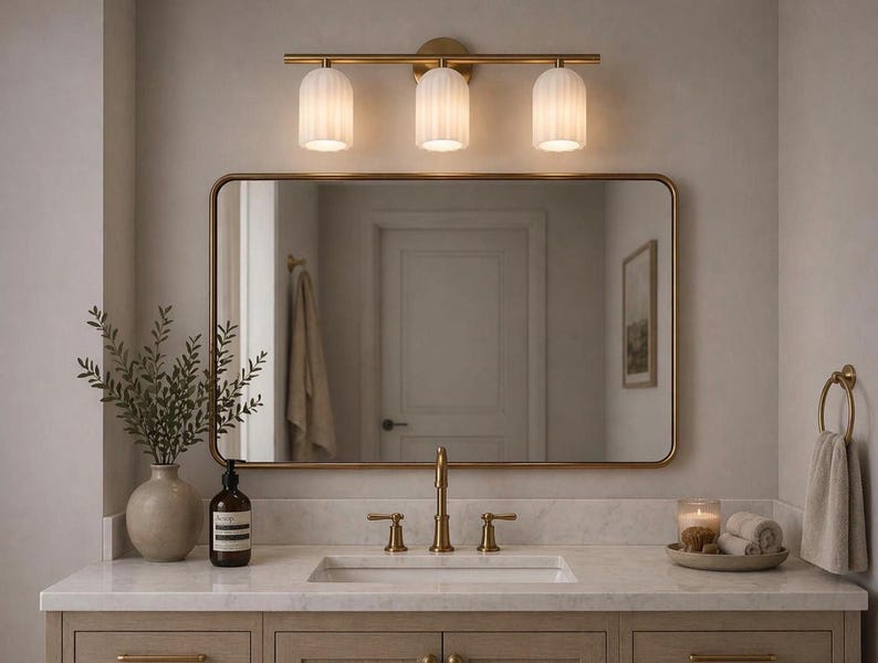 May include: A bathroom vanity with a gold-framed mirror, a three-light fixture, and a marble countertop. The vanity features a sink, faucet, and a decorative vase with greenery. A towel ring and a tray with candles and rolled towels are also visible.