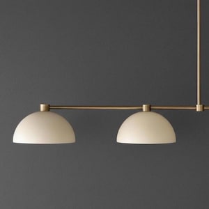 May include: A linear pendant light fixture featuring a horizontal brass rod and two matte off-white dome-shaped shades. The fixture is suspended by a vertical brass stem, showcasing a clean, symmetrical design with metallic and neutral tones.
