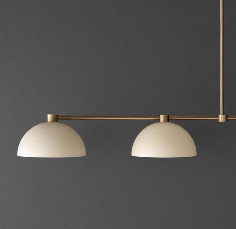 May include: A linear pendant light fixture featuring a horizontal brass rod and two matte off-white dome-shaped shades. The fixture is suspended by a vertical brass stem, showcasing a clean, symmetrical design with metallic and neutral tones.