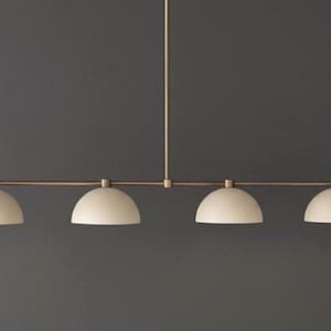 May include: A linear pendant light fixture featuring a thin, horizontal brass-toned metal rod suspended by a single vertical stem. Four matte off-white, dome-shaped shades are evenly spaced along the rod, each pointing downward to provide light.
