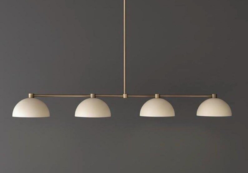May include: A linear pendant light fixture featuring a thin, horizontal brass-toned metal rod suspended by a single vertical stem. Four matte off-white, dome-shaped shades are evenly spaced along the rod, each pointing downward to provide light.