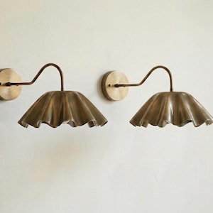 Op de afbeelding: Twee messing wandkandelaars met een gebogen arm en een geplooid, bloemblad-vormig kapje. De wandkandelaars zijn gemonteerd op ronde messing achterplaten. Het ontwerp is elegant en zou een verscheidenheid aan interieurstijlen aanvullen.