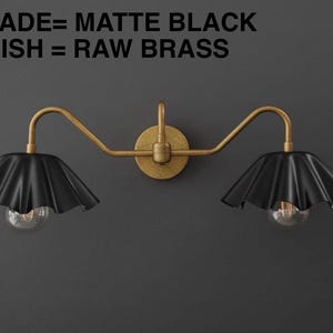 May include: A matte black and raw brass wall sconce with two lights. The sconce has a circular base and curved arms supporting the black, ruffled shades. The text in the image reads: SHADE= MATTE BLACK, FINISH = RAW BRASS.
