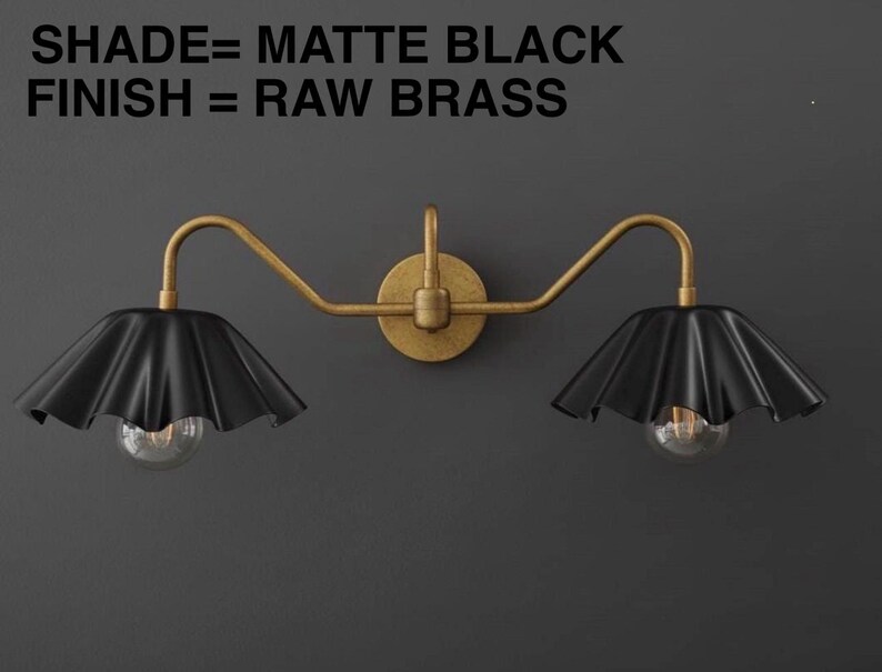 May include: A matte black and raw brass wall sconce with two lights. The sconce has a circular base and curved arms supporting the black, ruffled shades. The text in the image reads: SHADE= MATTE BLACK, FINISH = RAW BRASS.