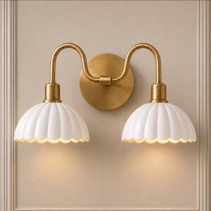 May include: A double wall sconce featuring a circular brass backplate and two curved brass arms extending to support two white, scalloped, dome-shaped shades. The fixture emits a soft downward light from each of the two white fluted lamp shades.