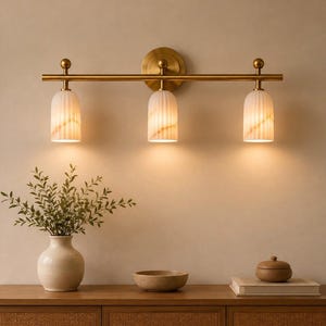 May include: A three-light wall sconce featuring a horizontal brass bar mounted to a circular backplate. Three downward-facing, fluted white glass shades are attached to the bar, each emitting a warm glow from the internal light source.